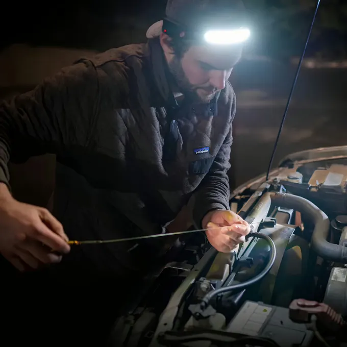 Mechanic leaning into an engine bay with both hands on the parts, lit by a headlamp