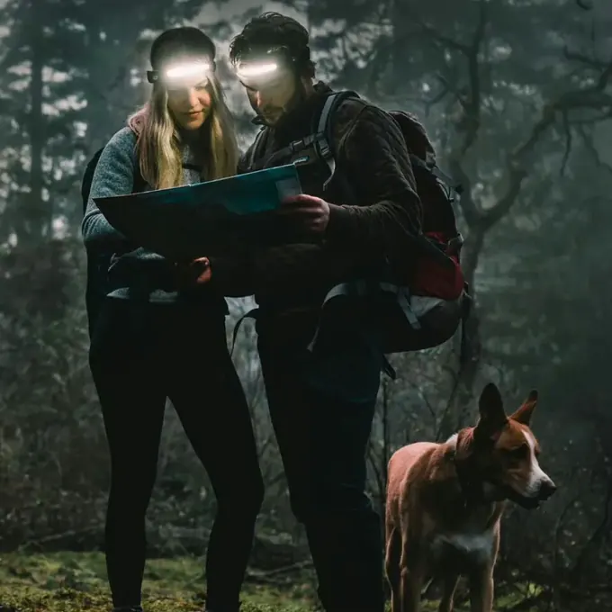 Two hikers wearing lit headlamps checking a paper map in a dark forest with their dog beside them