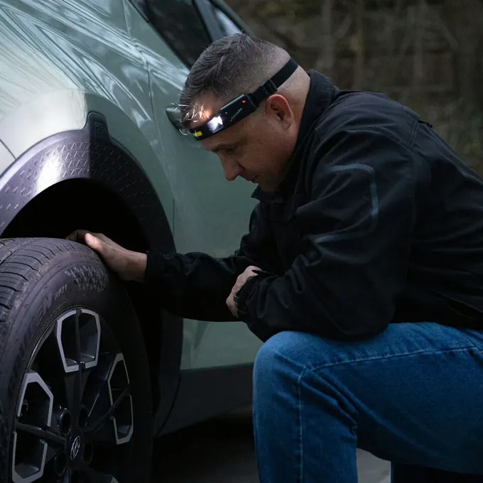 Man kneeling beside a car at night checking a tire by the light of his headlamp
